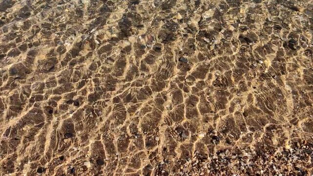 gentle rippling water over shallow sandy seabed with pebbles and shells, sunlit caustics and reflections creating tranquil, warm blue patterns evoking calm summer seaside vibes.