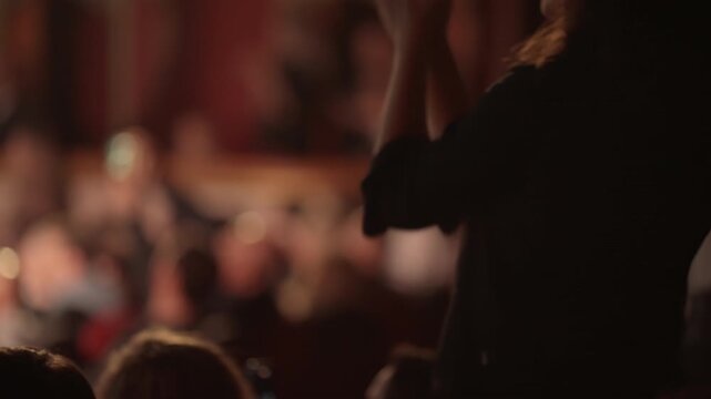 Close-up video of an audience member, seen from behind, enthusiastically clapping their hands in appreciation at a live event, concert, or performance. Blurred background of other attendees. Warm,