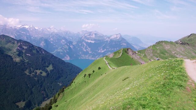 Drone aerial of hikers along Stoos Ridge in the Swiss Alps, overlooking Lake Lucerne, with lush green slopes and snow-capped peaks under a clear summer sky.