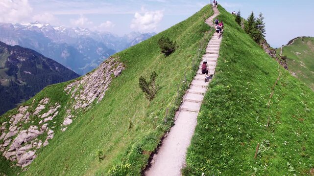 Mountain ridge hike, narrow alpine trail with steps, hikers on a grassy summit, dramatic Switzerland Alps scenery, summer adventure, aerial landscape travel footage.