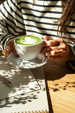 Woman hands holding cup of green matcha latte at sunlit cafe table with notebook, aesthetic morning routine and mindfulness concept