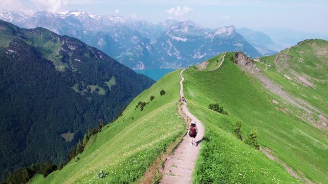 Aerial 4K drone view of hikers walking along the grassy Stoos Ridge in Switzerland, with sweeping alpine mountains, deep valleys, and clear summer skies creating a stunning scenic panorama.