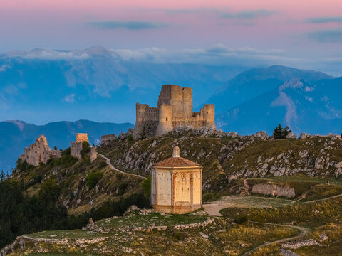 Aerial view of Rocca Calascio fortress and Santa Maria della Pieta chapel on a rocky ridge with the Apennine Mountains under a sunset sky in Calascio, Abruzzo, Italy.