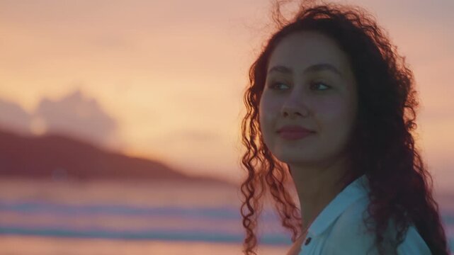 Curlyhaired woman with gentle smile overlooking ocean. Sunset scenery featuring woman on sandy shoreline with waves. Mixed woman with curly hair smiles softly against island silhouette at twilight