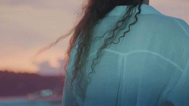 Woman watching sunset. Distant island scene highlighted by warm light. Curly haired woman gazes at sunset with windblown hair. Woman with curly hair observes sunset as wind blows her hair around