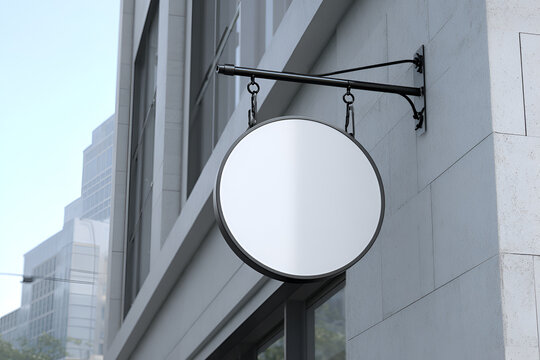 A blank, circular sign hangs from a decorative black bracket on a light-colored building wall, against a bright sky