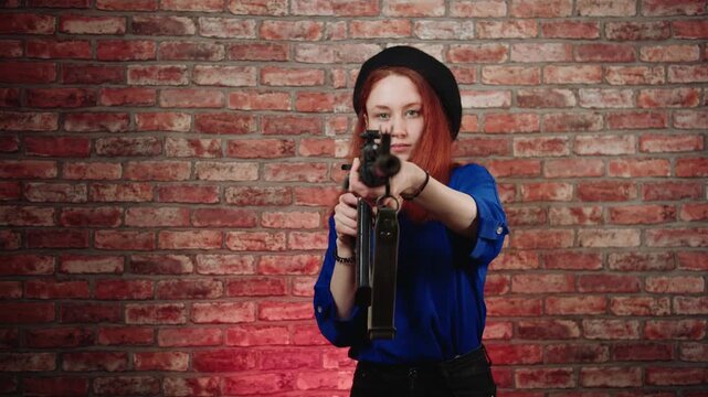 A portrait of a fighting caucasian young redhead woman in a blue blouse and black beret, aims a machine gun at the camera and fires with recoil against a brick wall. Kalashnikov assault rifle