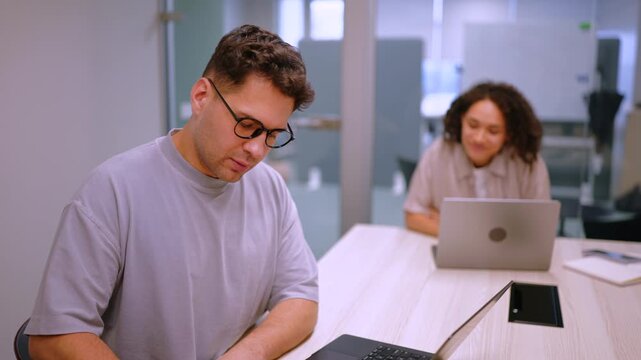 Man with glasses takes notes near laptop while female colleague types on computer across table. Contemporary office interior highlights efficiency, cooperation and structured business workflow.