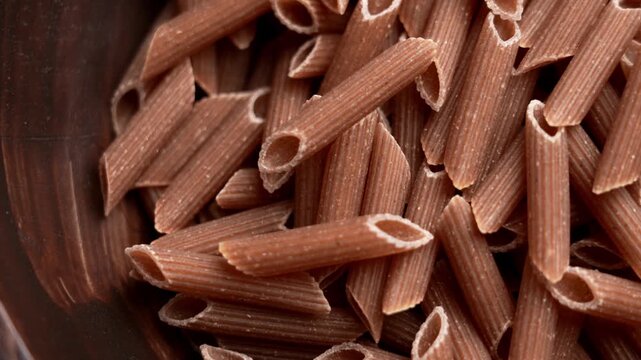 Raw wholemeal penne rigate pasta with rough ridged texture. Brown macaroni tubes scattered in a dark wooden bowl. Rotation. Top view