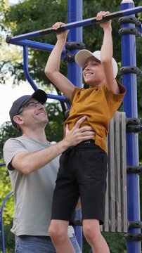 Child enjoying outdoor playground activity. Father helps son practice pull-ups in vertical position. Father and son are involved in sports. Joyful moments capture family bonding and healthy lifestyle.