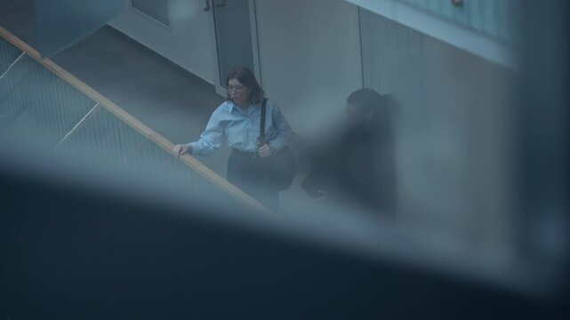 Tilt down shot of young Caucasian woman and biracial man in smart casual clothes talking while standing in modern business center leaning on handrail, seen from elevator lift