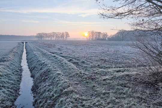 View of a serene winter landscape where the rising sun casts a golden glow over frost-covered fields and a tranquil waterway, Echten, Drenthe, Netherlands.