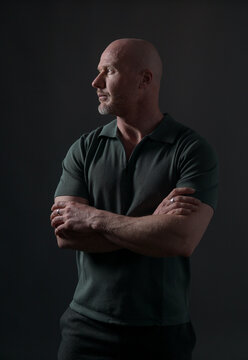 Confident mature bald man in dark green polo shirt looking aside on dark background, studio portrait with dramatic lighting