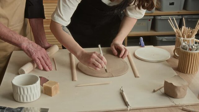 High angle shot of unrecognizable woman marking outline on flat, oval piece of clay while eldery craftswoman instructing her in modern pottery studio