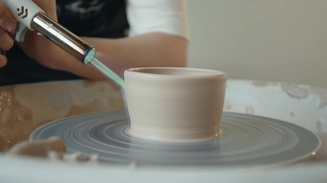 Close up shot of unrecognizable woman holding gas torch, directing it at small, unglazed ceramic cup on spinning potters wheel