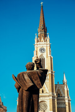 View of a bronze statue stands in quiet contemplation before the towering cathedral spire reaching up into the clear blue sky, Sad, Vojvodina, Serbia.