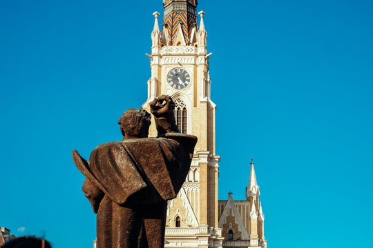 View of a statue's back against the towering, ornate clock tower under a bright blue sky, a serene blend of art and architecture, Sad, Vojvodina, Serbia.