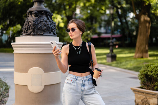 stylish woman with short haircut walking in street with coffee wearing black top, jeans and sunglasses accessories