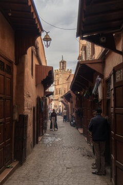 Marrakesh, Morocco - 13 November 2023: View of the bustling alleyways with the Koutoubia Mosque minaret rising in the distance, framed by aged, terracotta buildings.
