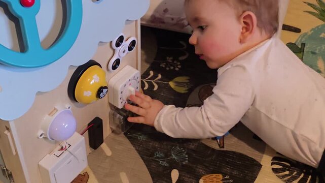 Curious baby lying on a colorful play mat and reaching out to play with various sensory elements on a wooden busy board at home indoors.