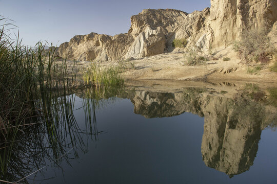 View of serene waters reflecting rugged cliffs and sparse vegetation under a clear sky, capturing the arid beauty of Hingol National Park, Hingol National Park, Balochistan, Pakistan.
