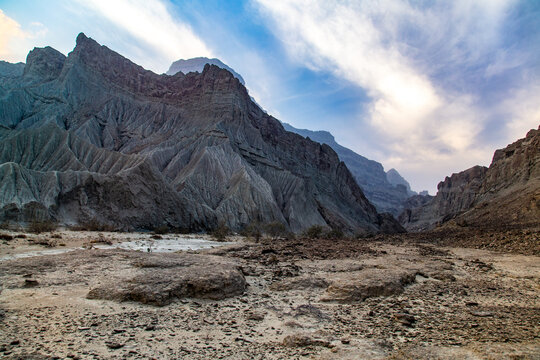 View of rugged, gray mountains rise against a blue sky streaked with white clouds, casting shadows on the arid landscape below, Hingol National Park, Balochistan, Pakistan.