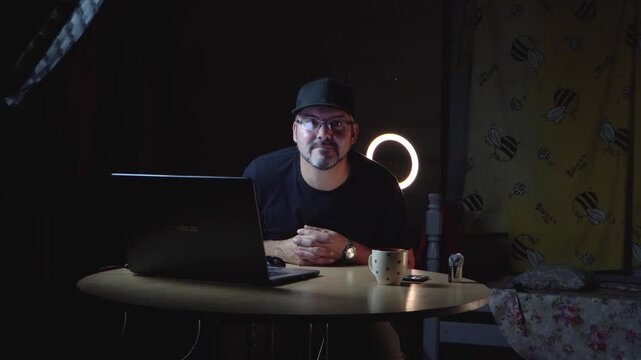 A man in black clothes sits at a table. He wears a baseball cap and glasses. He works remotely. Freelancer. Night. The lights are on. There's a laptop on the table. A ceramic mug. Home interior. Stora