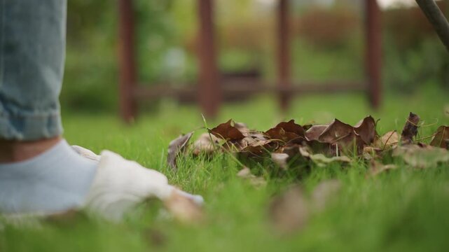 Closeup socked feet raking leaves, late autumn backyard scene with denim jeans, soft green grass, small leaf pile near wooden fence, gentle slow motion, domestic chore vibe, calm reflective