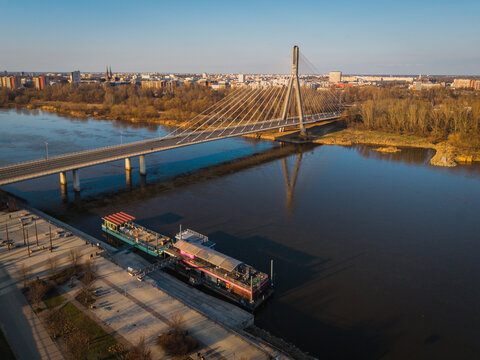 Aerial view of the Slasko-Dabrowski Bridge casting a stark shadow over the Vistula River, with the city skyline shimmering in the distance, Warsaw, Poland.