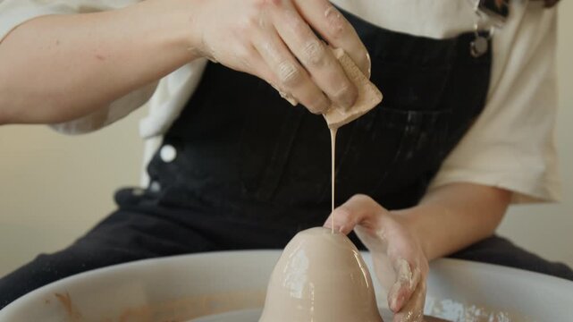 Close up tilt up shot of unrecognizable female hands in clay, pouring water with sponge and shaping cone-shaped object on spinning potters wheel