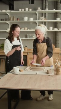 Vertical shot of elderly craftswoman and apprentice cutting shapes with stencil, molding clay in modern studio