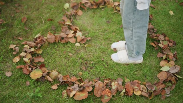 Person standing inside heartshaped leaf circle on green lawn wearing gray loungewear and white slippers, autumn leaves arranged into heart, cozy reflective mood, soft natural light, closeup of lower