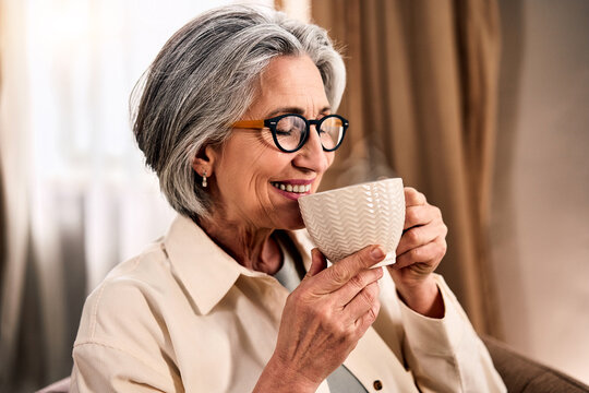 Smiling older woman with gray hair and glasses savoring a hot beverage from a patterned mug in a cozy indoor setting.