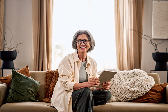 Smiling older woman with gray hair and glasses sitting on a cozy sofa holding a tablet in warm cozy living room. Comfort resting and cozy at home with digital gadget.