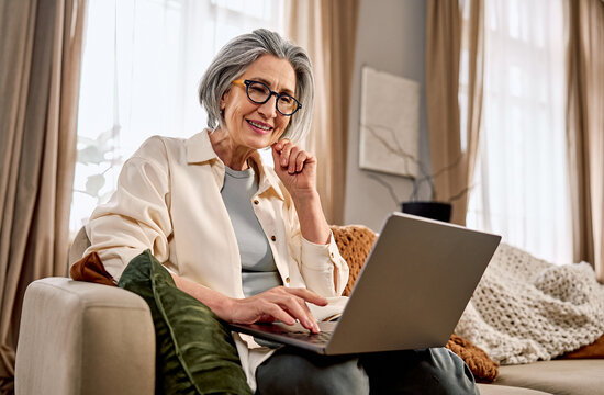 Beautiful older woman working on a laptop on a sofa in a cozy, bright living room.