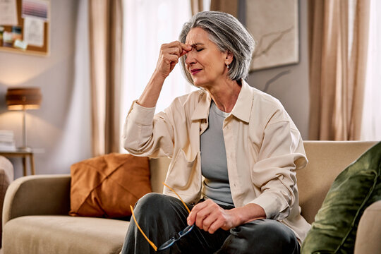 An older woman is sitting on a sofa, having taken off her glasses, experiencing a headache and pressure, dizziness, stress, and discomfort, holding her hand to her forehead with her eyes closed.