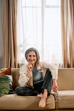 A happy, calm, positive, and beautiful senior woman in comfortable home clothes sits on a cozy sofa, holding glasses in her hand, looking at the camera and smiling. Vertical photo, relaxing at home.
