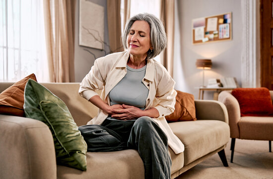 A senior woman is suffering from stomach pain while sitting on a sofa in the living room.