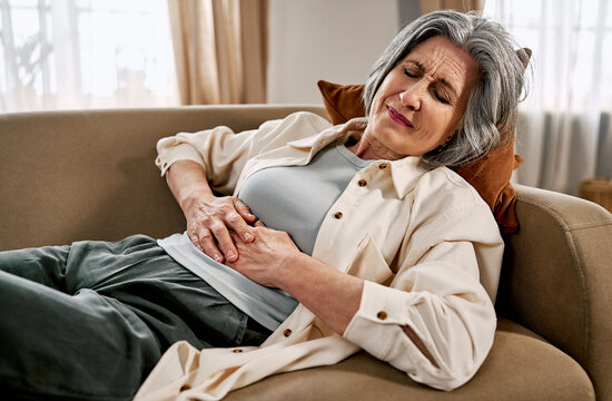 An older woman is lying on a sofa with her eyes closed, suffering from stomach pain. She feels discomfort and pain.