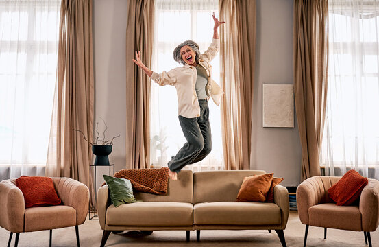 A happy senior woman is joyfully jumping on a sofa in her own apartment, in a cozy interior with a sofa and armchairs. A new homeowner celebrates the purchase of her property.