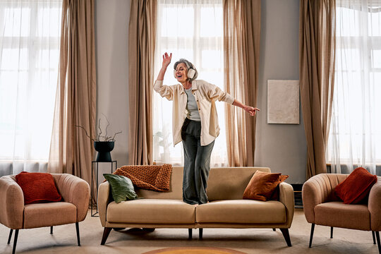 A happy woman in stylish home clothes is listening to music and dancing on a beautiful, comfortable sofa in a bright, spacious living room.