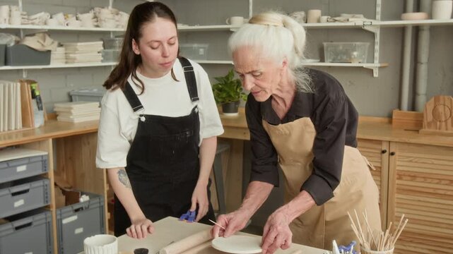 Young woman shaping clay using stencil after eldery craftswoman teaching her at workbench in modern pottery studio