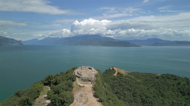 Fast drone approach to Rocca di Manerba del Garda ruins, descending toward the stone terrace and viewpoint cross as Lake Garda spreads blue behind summer clouds