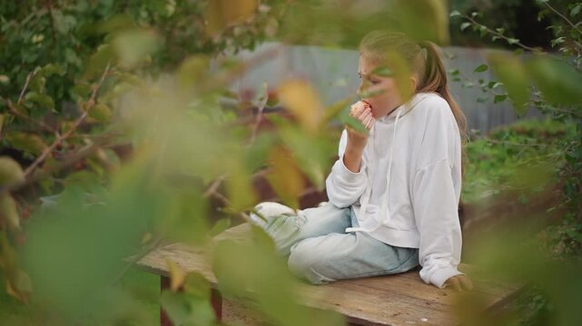 Caucasian girl through leafy branches peeking while eating apple on wooden bench, hoodie and jeans with ponytail, blurred foreground foliage creating dreamy voyeur perspective, quiet picnic mood,