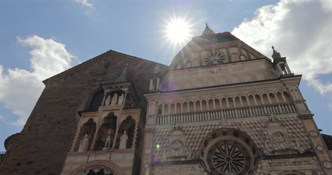 Sunlit upward view in Bergamo Italy starts on Duomo di Bergamo, lateral pan reveals Colleoni Chapel carved marble and finally frames the baptistery roof angel in open blue sky