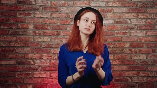 A portrait of a confident caucasian young redhead woman in a blue blouse and black beret, sprays perfume, rubbing the eau de toilette onto her skin against brick wall. Female fragrance and aroma