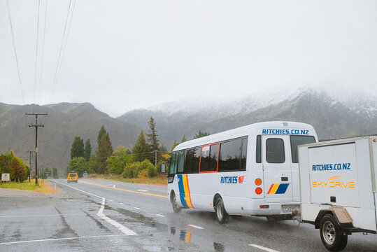 South Island, New Zealand: White SkyDrive passenger shuttle with trailer travels on wet mountain road during rain storm near Southern Alps.