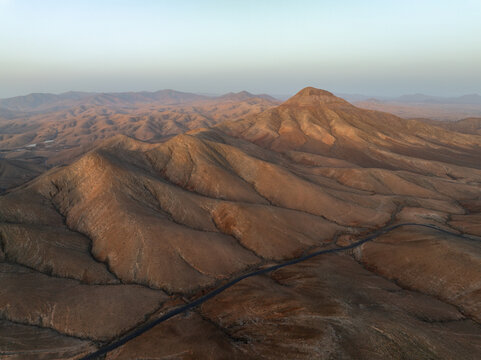 Aerial view of sun-kissed ridges and valleys carve a breathtaking tapestry across the arid landscape, punctuated by a solitary road, Pajara, Canarias, Spain.