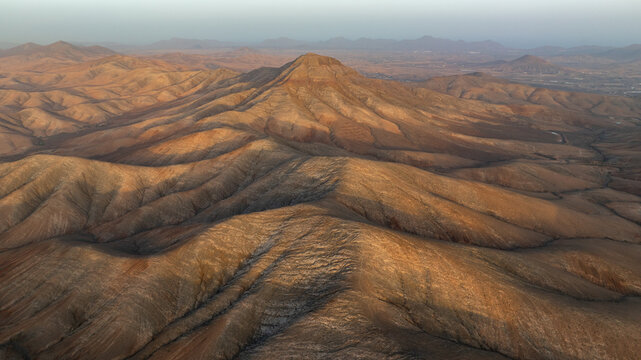 Aerial view of sun-kissed mountains casting long shadows across the rugged terrain, creating a dramatic landscape of warm hues and cool contrasts, Pajara, Spain.
