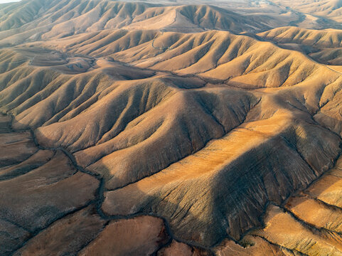 Aerial view of the undulating, sun-kissed mountains casting deep shadows across the arid landscape, creating a mesmerizing play of light and form, Pajara, Canarias, Spain.